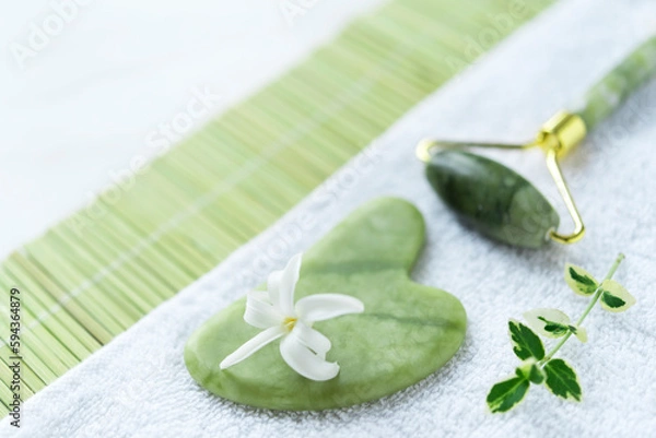 Fototapeta Spa composition of green jade Gua sha and roller, white flower, small branch of green leaves on the white towel and green bamboo napkin. Freshness. Flat lay 