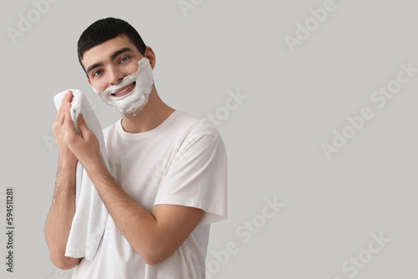 Fototapeta Young man wiping shaving foam from face against light background