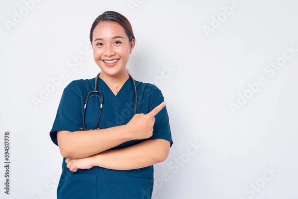 Fototapeta Smiling young Asian female professional nurse working wearing a blue uniform standing confident while pointing finger at empty copy space isolated on white background. Healthcare medicine concept
