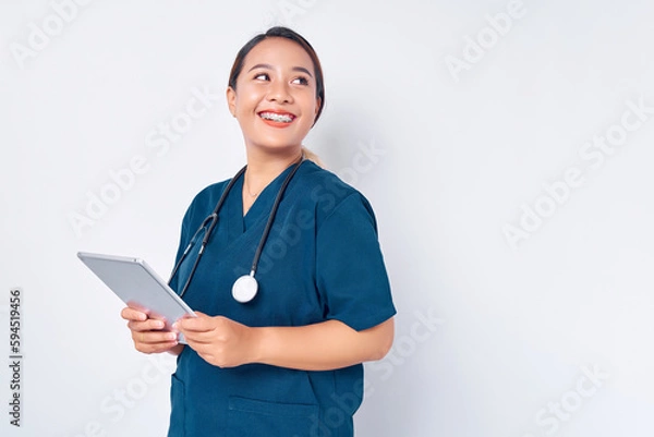 Fototapeta Smiling young Asian woman nurse working wearing a blue uniform using digital tablet to practice medicine and healthcare isolated on white background. Healthcare medicine concept