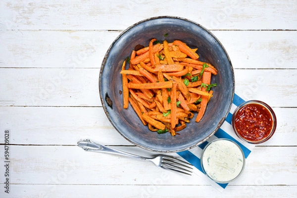 Fototapeta Homemade carrot chips in old antique bowl, silver fork, hot chilli and garlic sauce on white wooden desk. Top view.