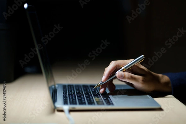 Fototapeta Business planning, E learning concept. Man hand with pen working on laptop computer at home office, close up, dark room. Student lecture while attending online course, information searching.