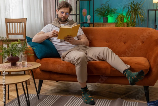 Fototapeta Young man relaxing interesting book, turning pages smiling enjoying literature, taking a rest on comfortable couch. Portrait of peaceful cheerful Caucasian guy at home apartment living room on sofa