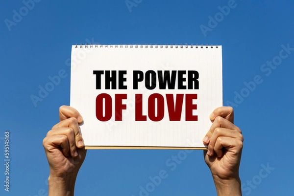 Fototapeta The power of love text on notebook paper held by 2 hands with isolated blue sky background. This message can be used as business concept about love.