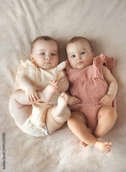 Fototapeta Vetical photo of two sisters baby girls in similar cotton clothes lying on bed. Fraternal twins .