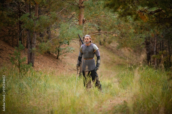Fototapeta an old warrior in chain mail with a sword is looking for prey in the forest.