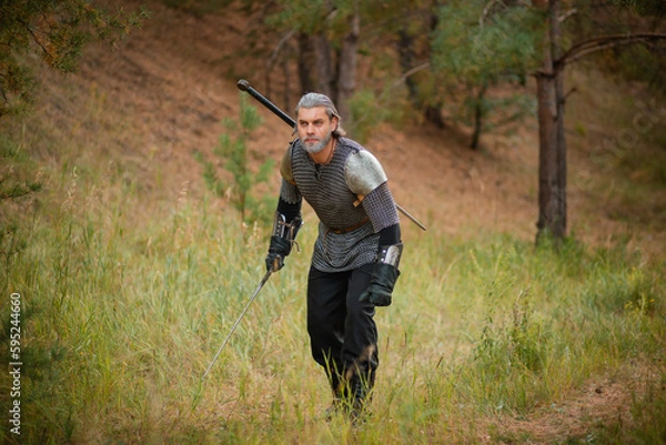 Fototapeta an old warrior in chain mail with a sword is looking for prey in the forest.