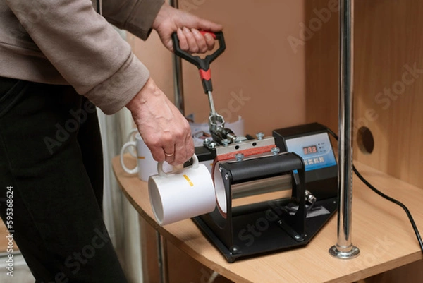 Fototapeta A woman at home learns to make souvenir cups on a heat press, a paper with a printout of an image for applying to a cup is attached to the cup with heat-resistant tape