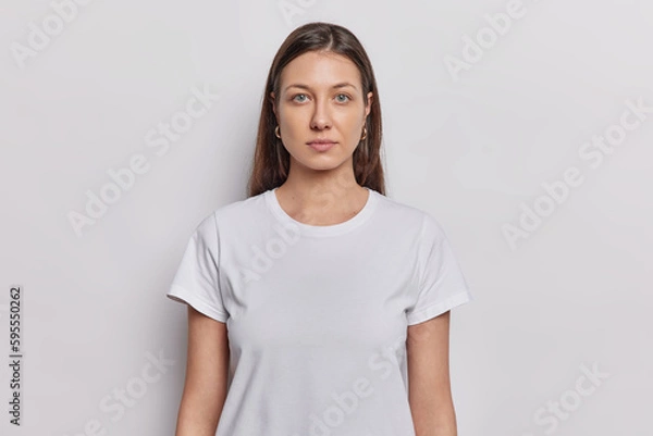 Fototapeta Waist up shot of nice looking woman has dark hair dressed in basic t shirt keeps arms down looks directly at camera isolated on white background. Brunette female model poses in studio for making photo