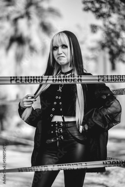 Obraz Dangerously stylish: young woman in goth accessories rocks black outfit and poses with funny face behind a danger line in park (in black and white)