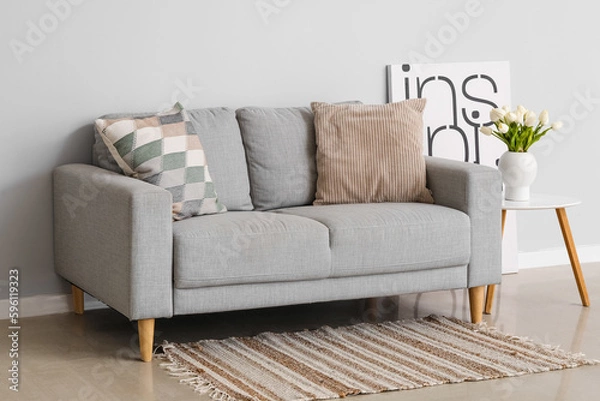 Fototapeta Cozy sofa with cushions and tulip flowers in vase on table near grey wall