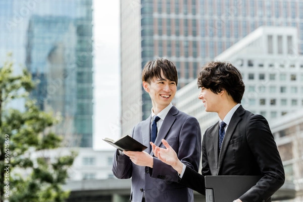 Obraz ビジネス街を歩きながら30代の先輩の言う事をメモする20代の後輩の新入社員2人のチームワーク