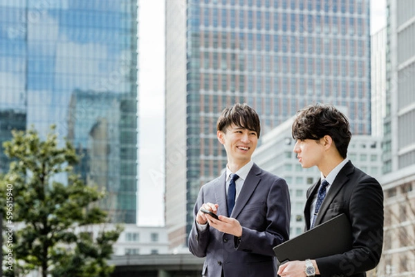 Obraz 高層ビルに囲まれた都会の屋外で楽しそうに会話をする20代から30代のスーツのビジネスマンの男性2人