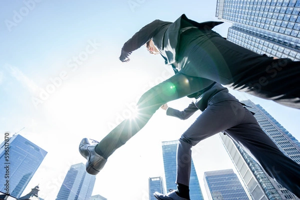 Obraz 青空の下ビルに囲まれた都会をジャンプで通り過ぎる2人のスーツのビジネスパーソンの男性