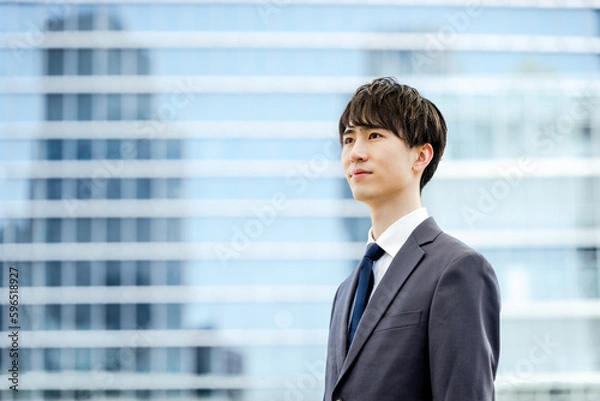 Obraz オフィスビルのガラスの建物の前で真面目な表情で遠くを見つめる20代のスーツを着た新入社員の男性
