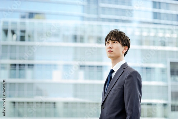 Obraz 高層ビルのガラスの壁面を背景に遠くを見つめる20代のスーツを着たビジネスマンの男性1人