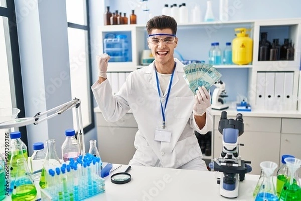 Fototapeta Young hispanic man working at scientist laboratory holding brazilian reals screaming proud, celebrating victory and success very excited with raised arm