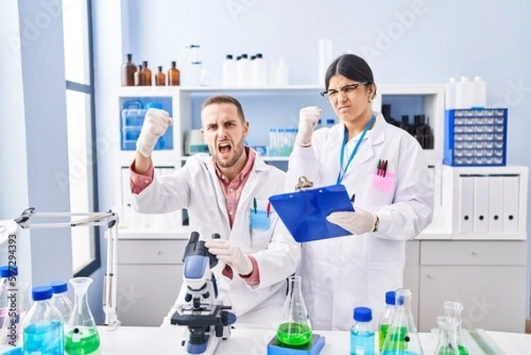 Fototapeta Two young people working at scientist laboratory annoyed and frustrated shouting with anger, yelling crazy with anger and hand raised