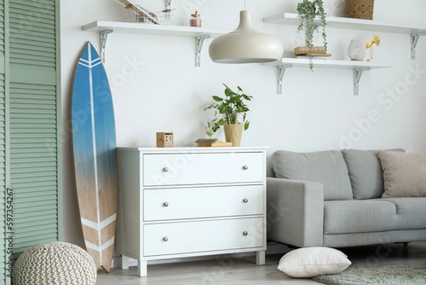 Fototapeta Interior of light living room with surfboard, commode and sofa