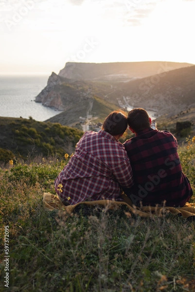 Fototapeta Travelers watch the sunset in the mountains.The couple meets the sunset in the mountains. Two travelers are sitting on the edge of a cliff and admiring the beautiful sunset. Panoramic view.