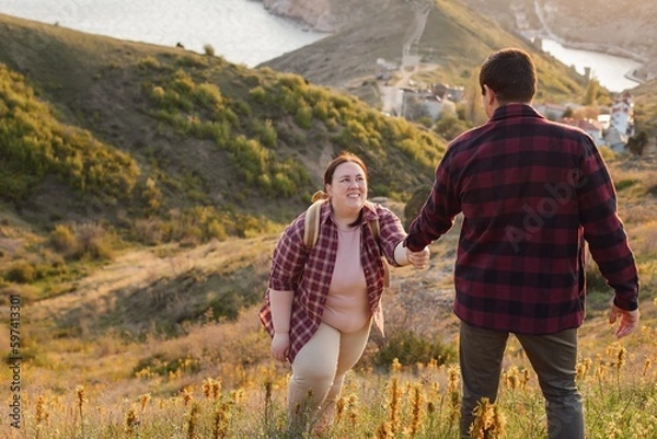 Obraz Tourists: A couple travels through the spring mountains. A man in a plaid shirt gives his hand to help a plump woman up the mountain. Walks in the open air. Travel and active life concept. Travel life