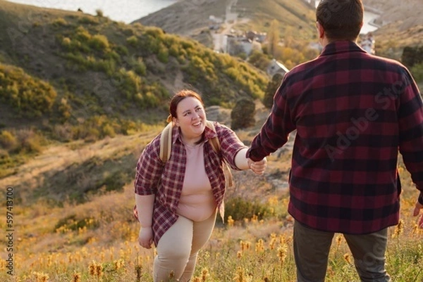 Fototapeta Tourists: A couple travels through the spring mountains. A man in a plaid shirt gives his hand to help a plump woman up the mountain. Walks in the open air. Travel and active life concept. Travel life