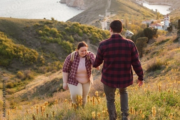 Obraz Tourists: A couple travels through the spring mountains. A man in a plaid shirt gives his hand to help a plump woman up the mountain. Walks in the open air. Travel and active life concept. Travel life