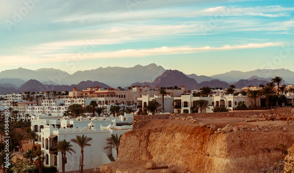 Fototapeta Imposing desert mountains provide a stunning backdrop to a cluster of white buildings, nestled amidst flourishing palm trees, capturing the essence of a tranquil desert oasis