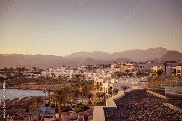 Fototapeta Imposing desert mountains provide a stunning backdrop to a cluster of white buildings, nestled amidst flourishing palm trees, capturing the essence of a tranquil desert oasis