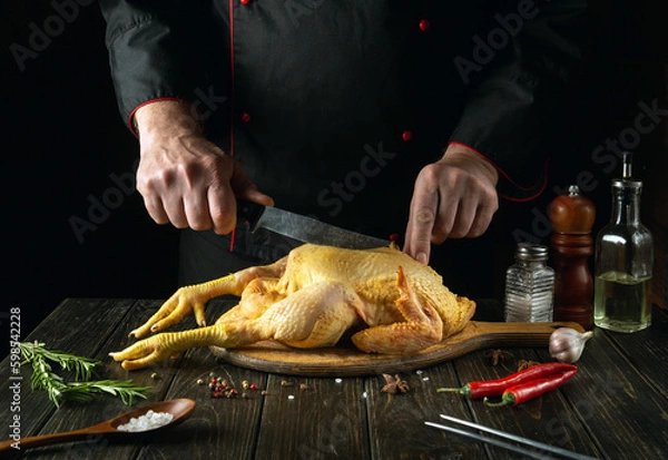 Fototapeta The chef prepares to cut a raw rooster with a knife on a kitchen cutting board. Cooking cockerel with fragrant spices and rosemary for lunch. Asian cuisine