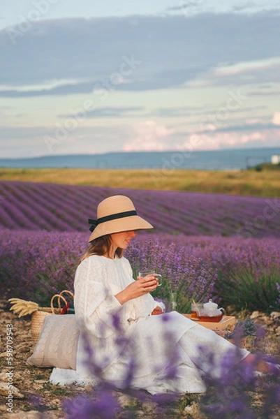 Obraz A woman in a white dress sits in the middle of a lavender field and holds fragrant tea in her hands. Blooming lavender. Picnic in Provence style.