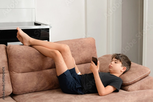Obraz The boy plays on the sofa in the living room. Living room kitchen with sofa. The child plays with gadgets. The boy spends his summer vacation playing games on his phone.