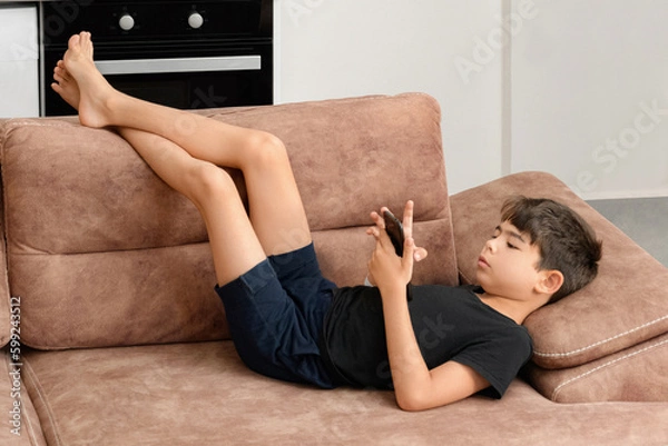 Obraz The boy plays on the sofa in the living room. Living room kitchen with sofa. The child plays with gadgets. The boy spends his summer vacation playing games on his phone.