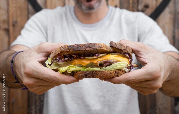Fototapeta View of a man holding a sandwich made with bread, grilled chicken, lettuce, sliced cabbage, cucumber and cheddar cheese.