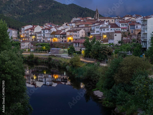 Fototapeta Night view of the town of Ribadavia in Orense, reflected with the lights in the Avia river, with the bell tower and white houses, in the background the mountain in summer of 2021 Spain