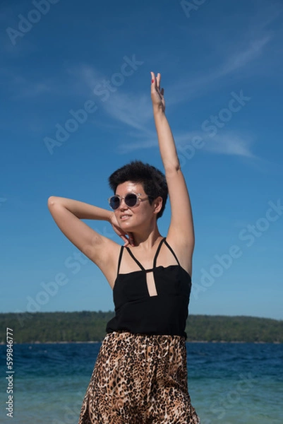Fototapeta Portrait of attractive girl standing near to the sea in summer vacation. Stylish pretty woman with short black hair wears in leopard pant having fun on the tropical beach with copy space.