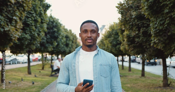 Fototapeta Front view of attractive smiling happy young black-skinned man in casual clothes which standing in the middle of urban alley with green trees and looking at camera on sunny day