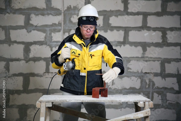 Fototapeta Locksmith in special clothes and goggles works in production. Worker. A man in a helmet and with a grinder in his hand. Industry. Builder. Metal processing with angle grinder