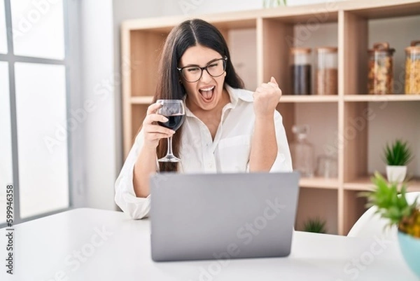 Fototapeta Young brunette woman doing video call with laptop drinking a glass of wine screaming proud, celebrating victory and success very excited with raised arm