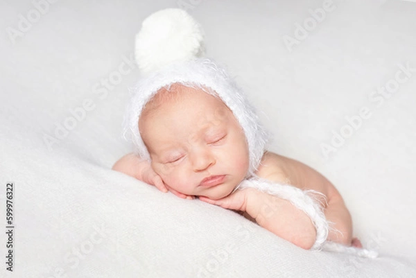Fototapeta a newborn baby in a white hat with a pom-pom sleeps on a white bedspread over white background with copy space. beautiful sleeping baby