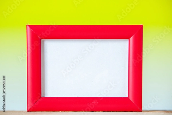 Obraz Close up view of red empty picture frame standing on table on yellow white mix color background with free space