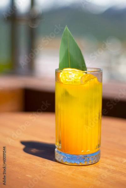 Fototapeta A glass with a cocktail, juice, lemonade on a table in the mountains. Rest. Relax. Fruity drink decorated with green leaf and orange chips.