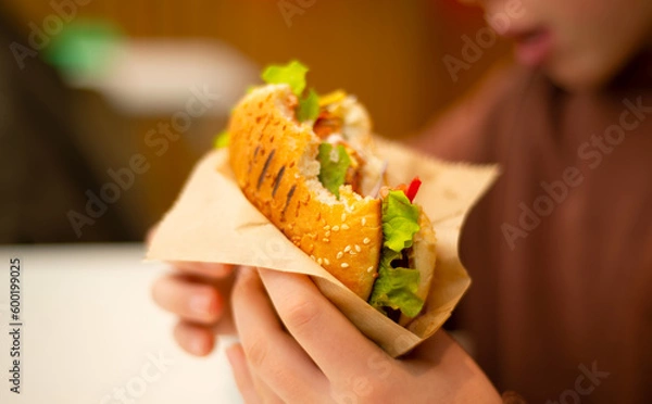 Fototapeta Burger with green lettuce in kraft paper in hands. Bitten piece. Grilled burger buns.