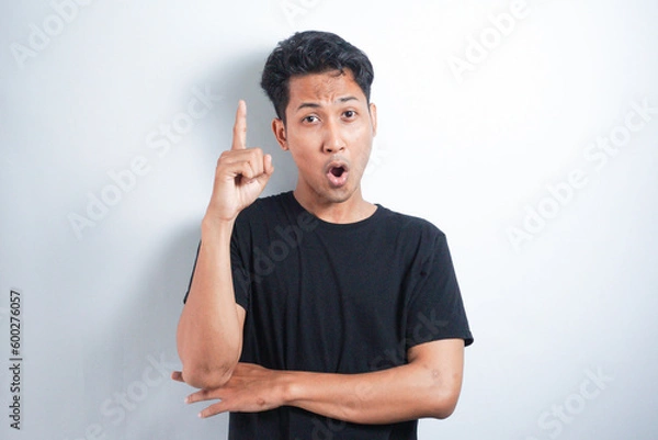 Fototapeta Young excited man in black t-shirt, pointing upwards with finger, having great idea, isolated on white background