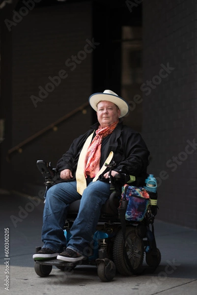 Fototapeta Outside of a stairwell, a man in a powerchair smiles confidently. Adding backstory, he is disabled and trans. His colorful scarf alludes to his LGBTQ identity.