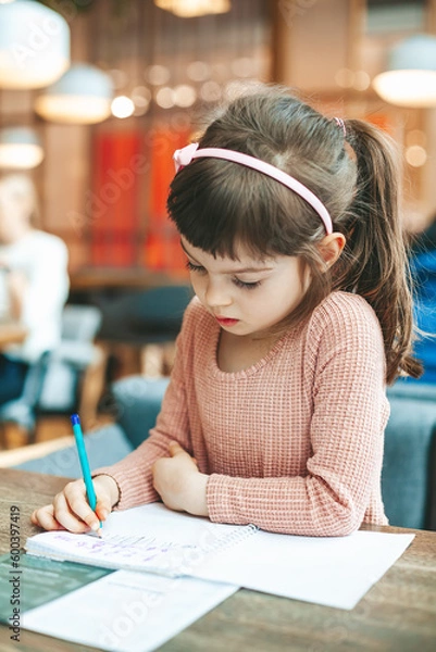 Fototapeta Cute little girl writing something in a notebook sitting in a cafe