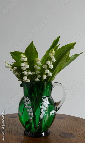 Fototapeta Freshly cut, sweetly scented Lily of the Valley (Convallaria majalis) in an Antique Victorian green glass jug with white enamelling of the  flowers on an oak table. Portrait image with space for text.