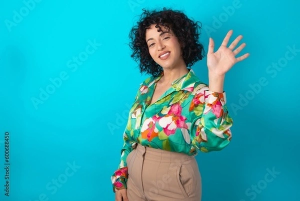 Fototapeta young arab woman wearing colorful shirt over blue background Waiving saying hello happy and smiling, friendly welcome gesture.