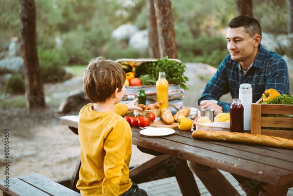 Fototapeta Father dad and school kid boy child having a picnic in the forest camping site with vegetables, juice, coffee, and croissants. Wooden crate with fresh organic veggies surrounded with bread baguettes