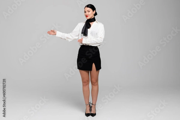 Fototapeta Portrait of an attractive young Stewardess. Charming, smiling air hostess in sensual uniform, gesturing, offering to come in. Isolated on white background.